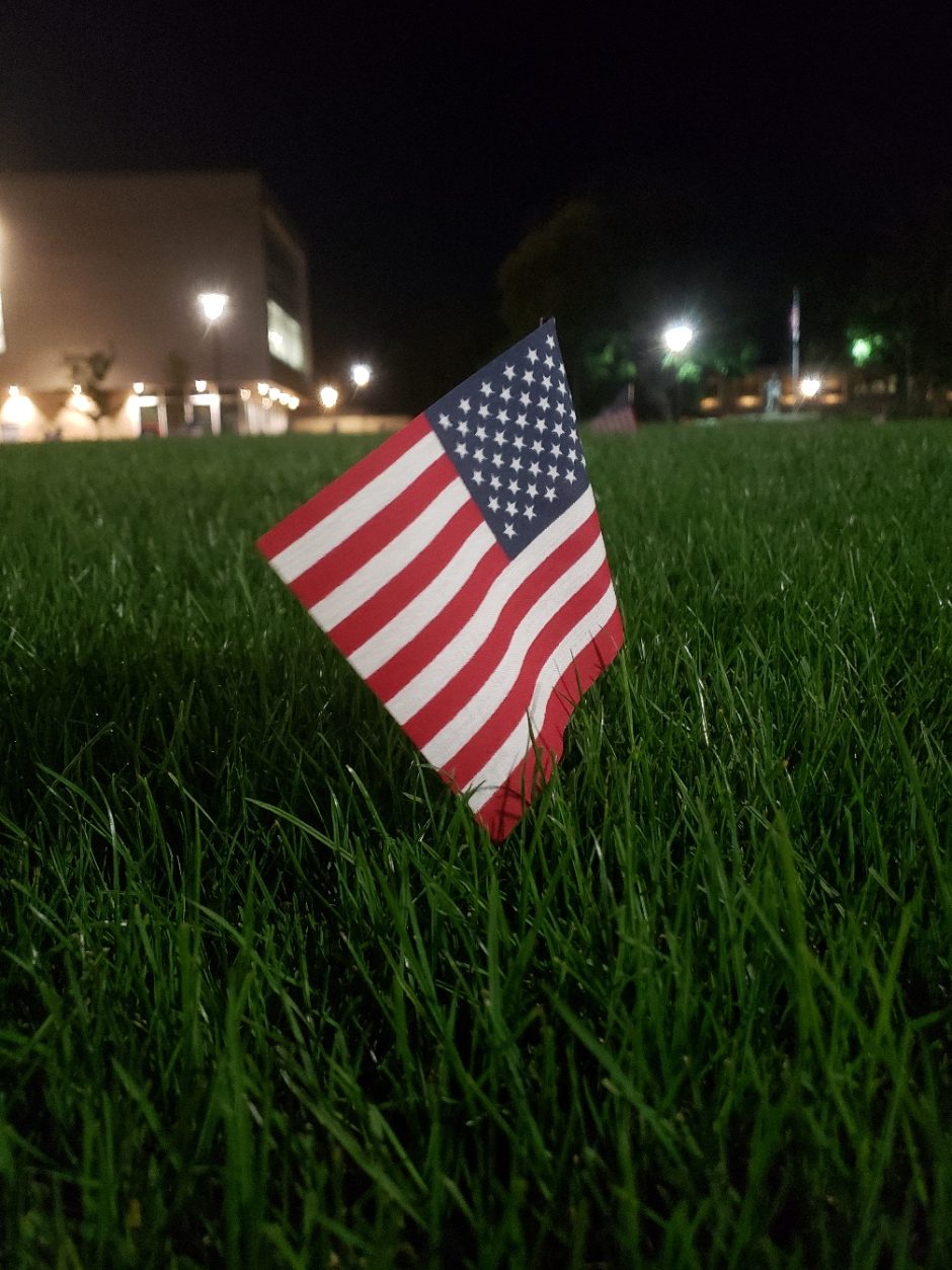American Flag planted in thr Grass at night with hazy lights in the distance